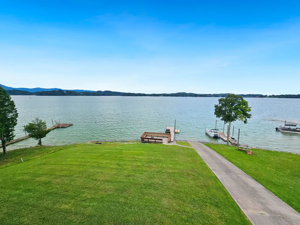 A broad view of the lakefront yard with a lush green lawn leading down to the water's edge, docks extending into the serene lake, and a clear pathway for easy access. The area is ideal for boating, fishing, and relaxing by the water, with sunrise light casting a tranquil mood.