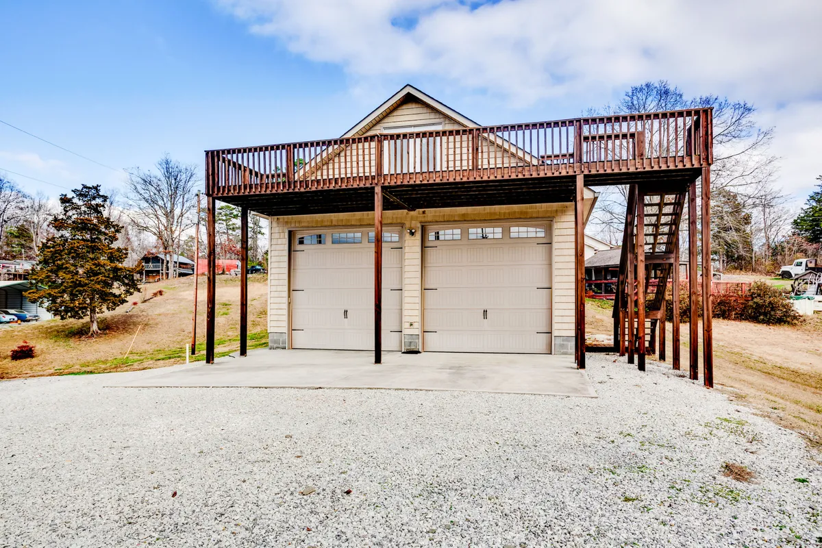 An exterior shot featuring the home’s welcoming entrance, wooden deck, and staircase, with two garage doors below and ample space for gathering. The deck is oriented toward stunning sunset views and surrounded by lush natural scenery.