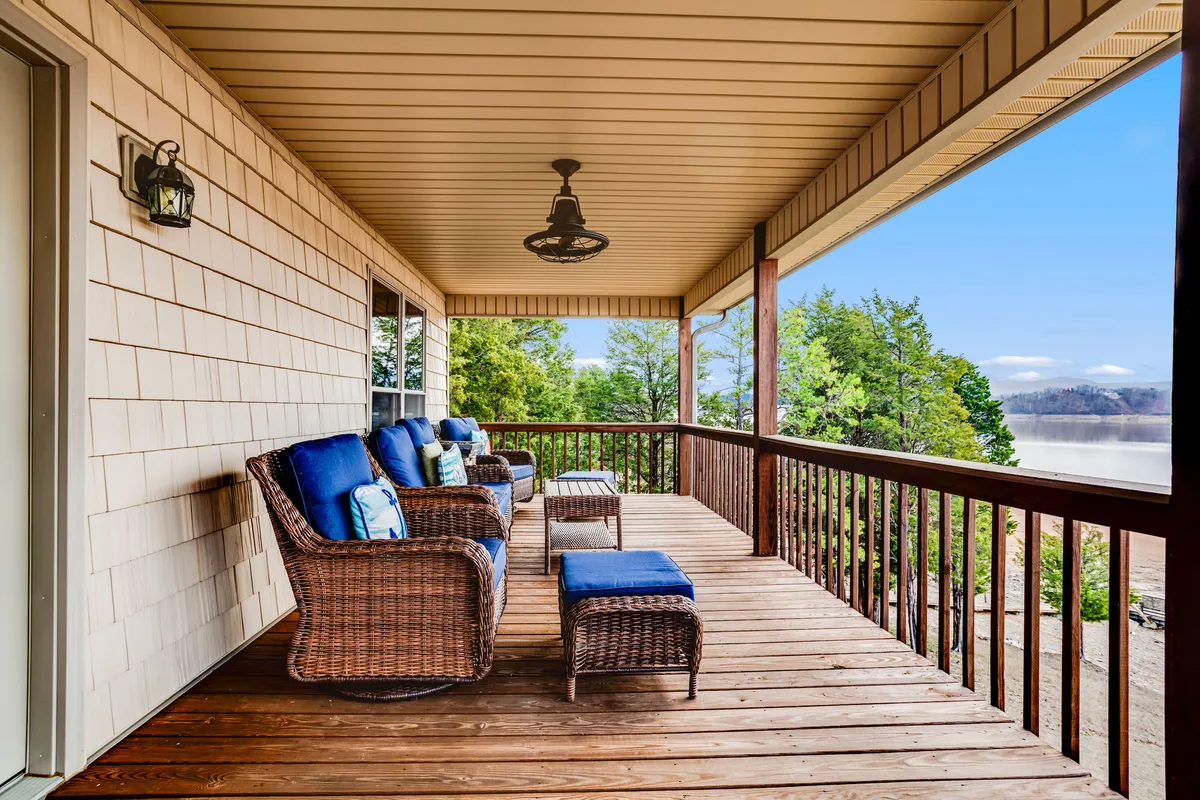 A serene view from a furnished exterior deck with wicker chairs featuring blue cushions, a wooden railing, and a sweeping vista of the lake and forested foothills. The tranquil setting is ideal for sunrise views, stargazing, and appreciating autumn foliage reflected in the water.