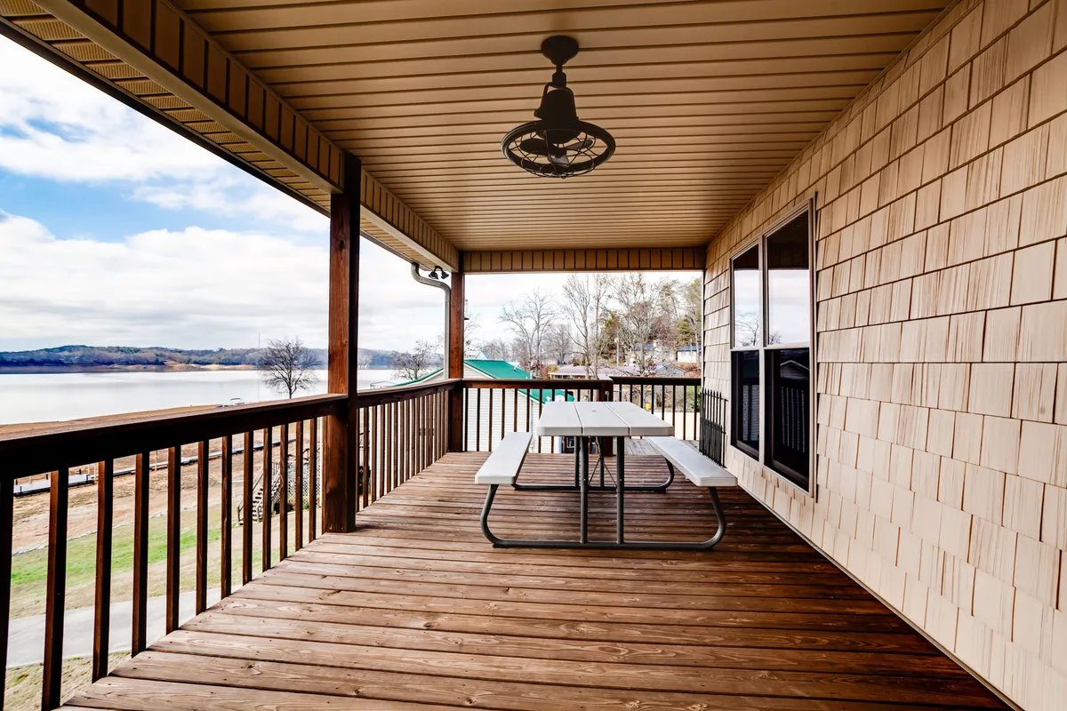 An outdoor scene showing a picnic table on a deck with a lake view and a wooden railing. The space is set up for outdoor dining, relaxation, and gathering, with the water just beyond. Decorative lighting suggests evenings spent socializing by the lake, echoing the fire pit gatherings and lakeside lifestyle described in the section.