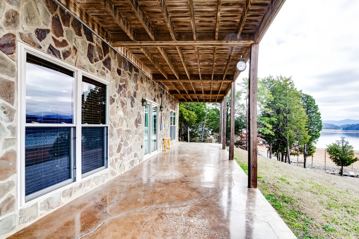 An exterior patio scene with a stone wall, wooden beams, and clear views of the lake. The area is set up for relaxation or social gatherings, evoking an inviting atmosphere for hosting barbecues or simply enjoying nature with a group.