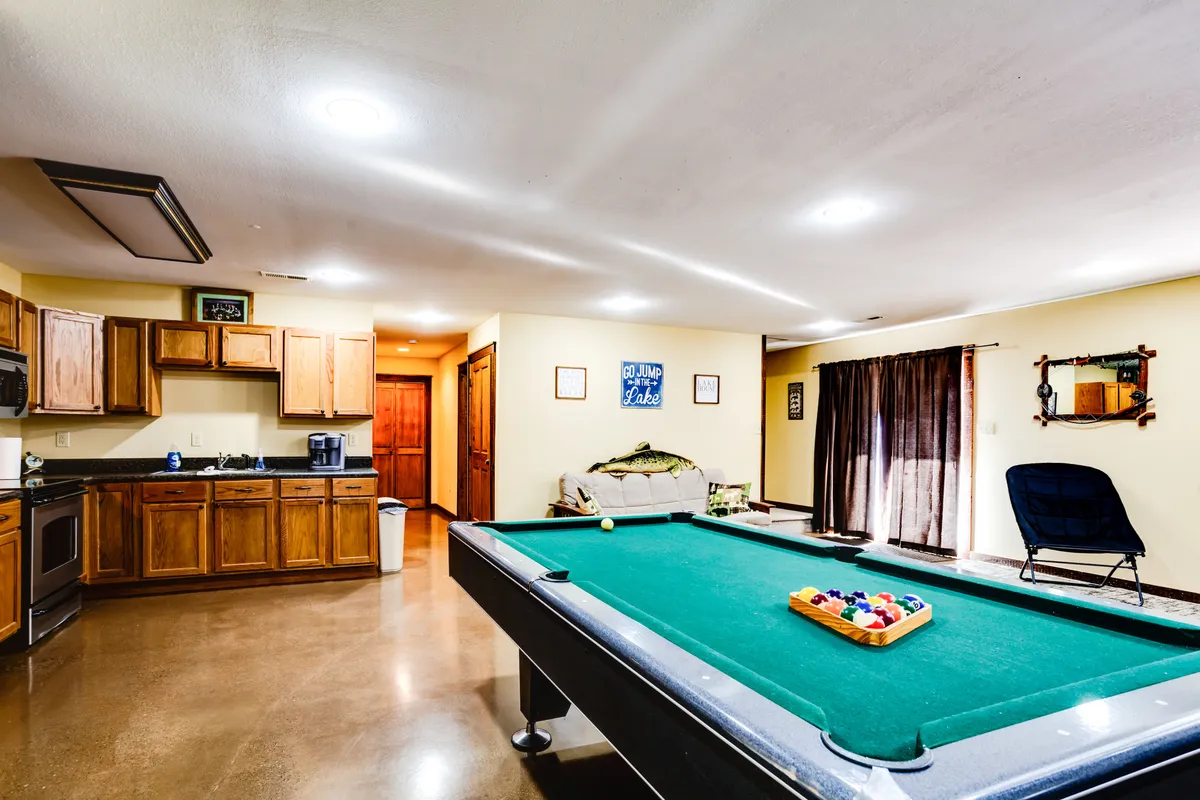 This image showcases a vibrant game room featuring a pool table at the center, a cozy sofa, a visible kitchen area with cabinetry and appliances, and a decorative mirror on the wall. The space appears casual and playful, perfect for socializing and group entertainment.