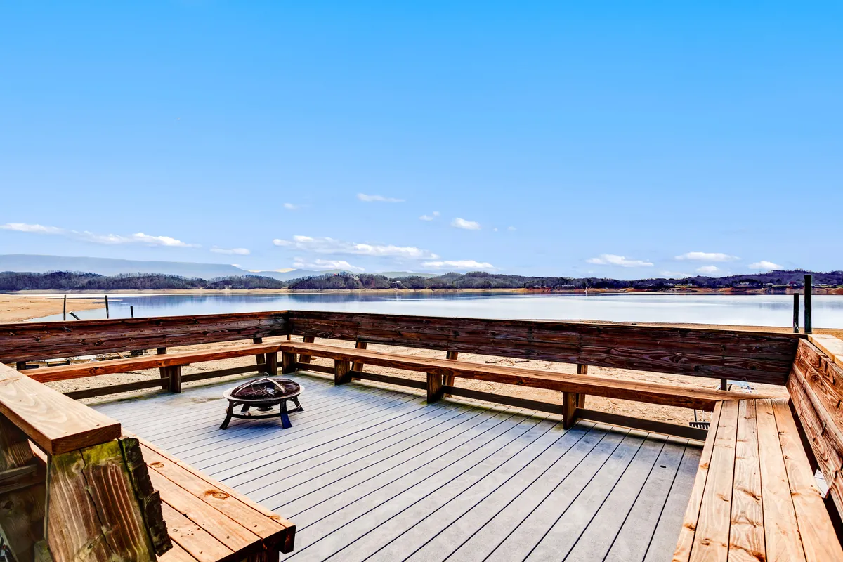 An exterior view of the furnished deck, overlooking the lake and mountains. Features a gas fire pit, wooden deck furniture, and an unobstructed view of the tranquil water and surrounding natural beauty—perfect for al fresco dining, morning coffee, or evening fireside chats.